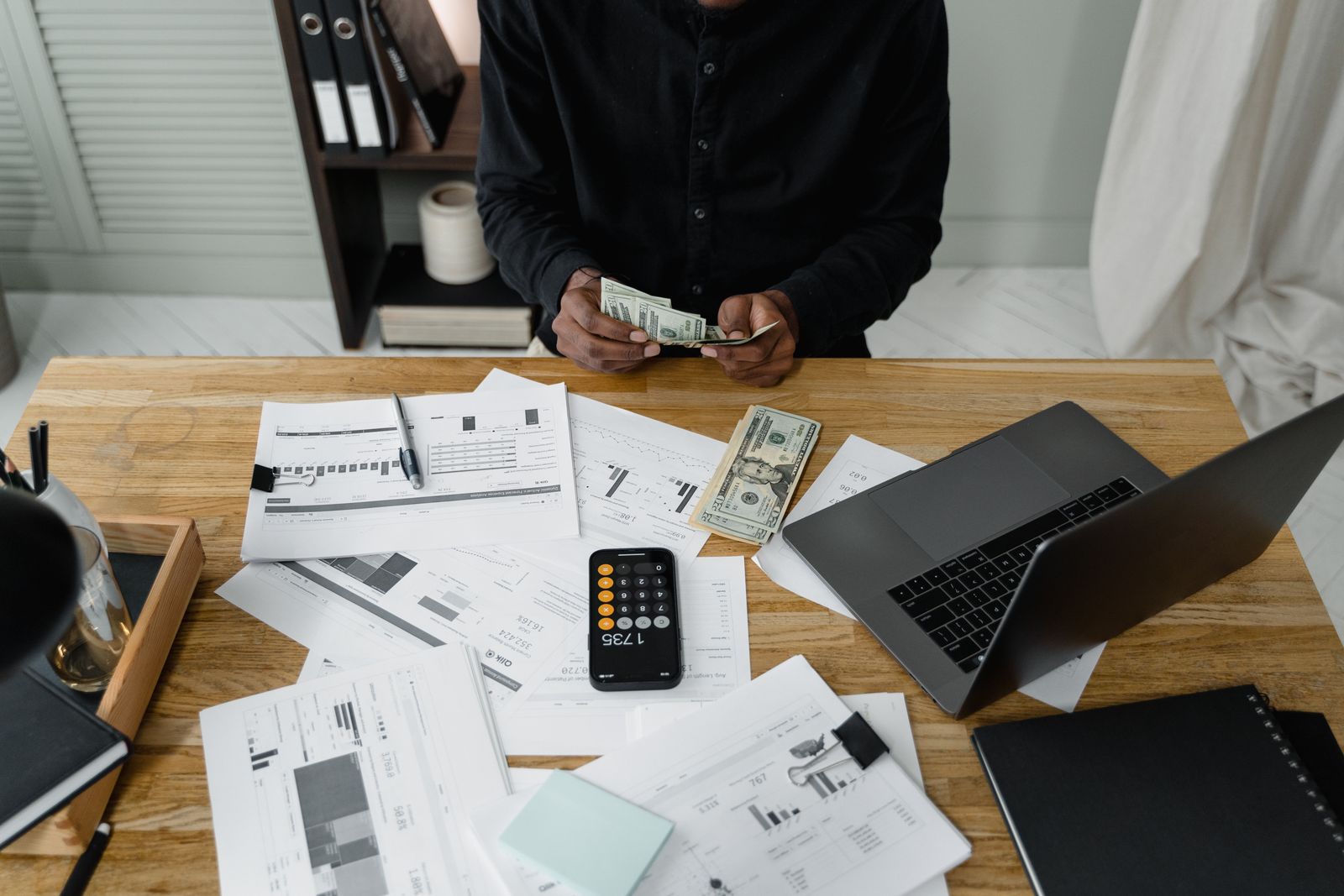 A person counts dollar bills at a desk with a laptop, papers, and calculator; young entrepreneurs personal finance